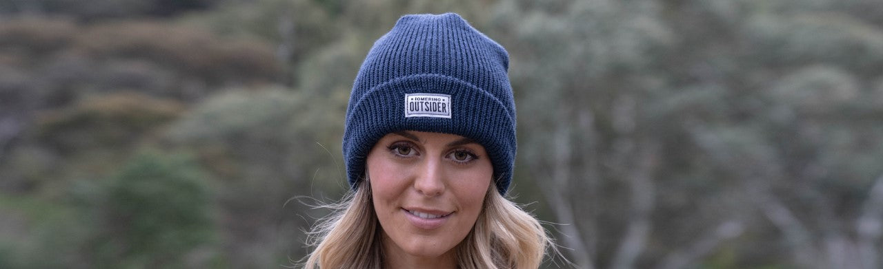 Image of a model's head, wearing ioMerino's Ribbed Beanie in Dark Blue, an out of focus background of trees behind her.