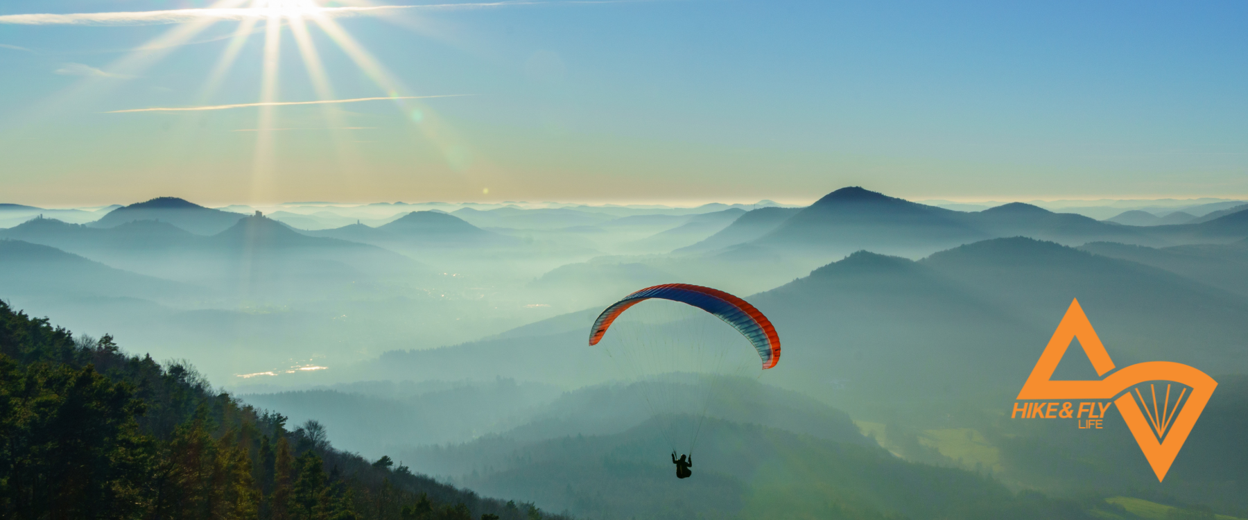 Image of a Hike & Fly participant, paragliding, misty mountains and clear skies in the background. In the bottom right corner is an orange logo for Hike & Fly Life.