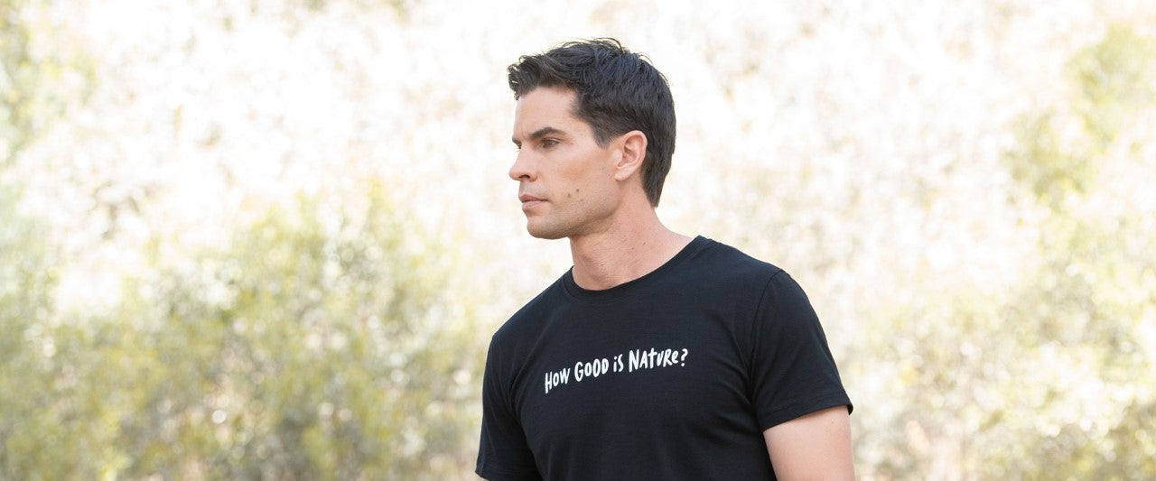 Image of a model wearing ioMerino's Men's Universal Tee in black, with 'How good is nature?' printed across the front in white text. Model is standing, facing left, standing in front of shrubbery.